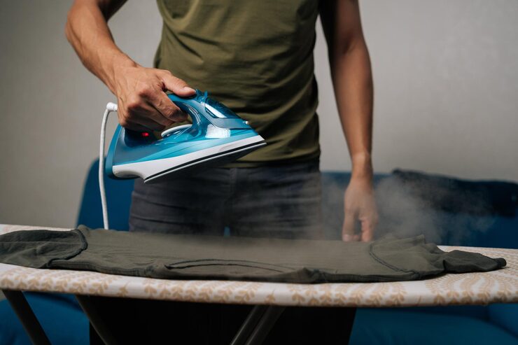 Person in a green shirt irons a shirt on an ironing board, steam rising from the fabric.